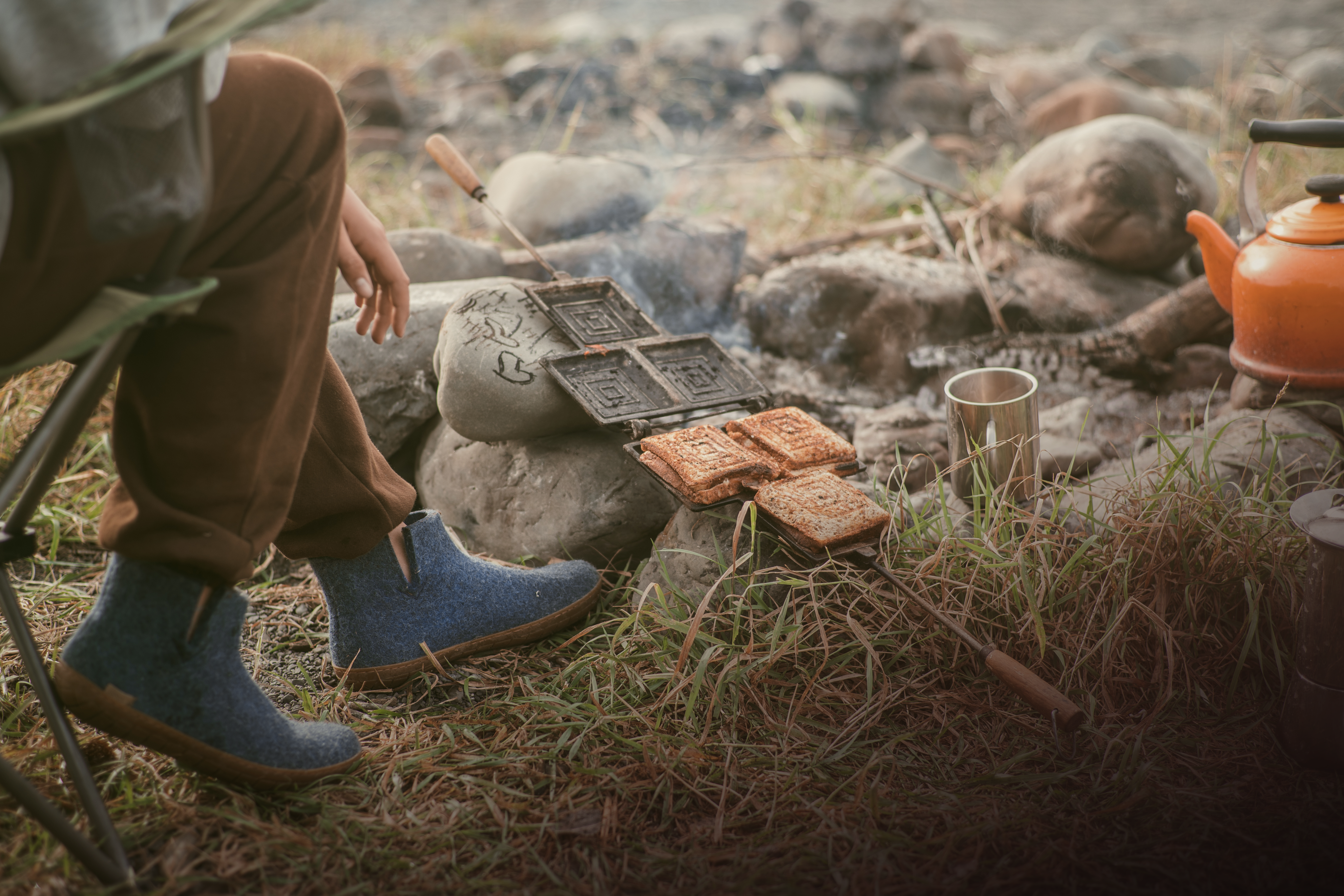 An outdoor, low-angle shot of a person sitting in a camping chair by a campfire, making toasted sandwiches in a cast-iron sandwich maker resting over the rocks, while wearing Denim, blue glerups boots with honey rubber soles.