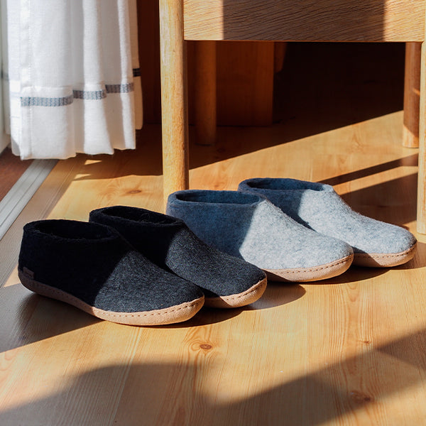 An indoor, high-angle shot of two pairs of felted glerups slip-on slippers sitting in a patch of bright morning sunlight on a light-colored hardwood floor near a wooden chair and a white striped curtain; one pair is Charcoal, black and the other is Grey, and both have leather soles shoe